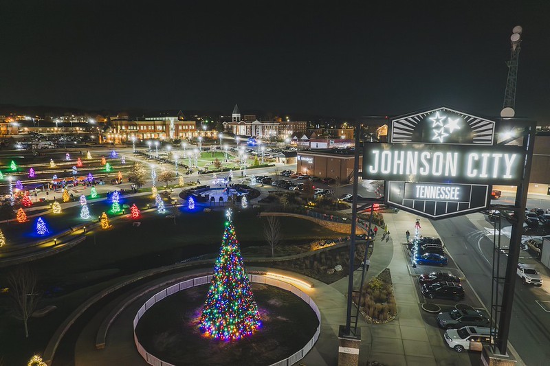 Elevated nighttime view of a downtown Johnson City park filled with glowing holiday trees and a large lit Christmas tree, with an illuminated Johnson City Tennessee sign at a past Jingle event